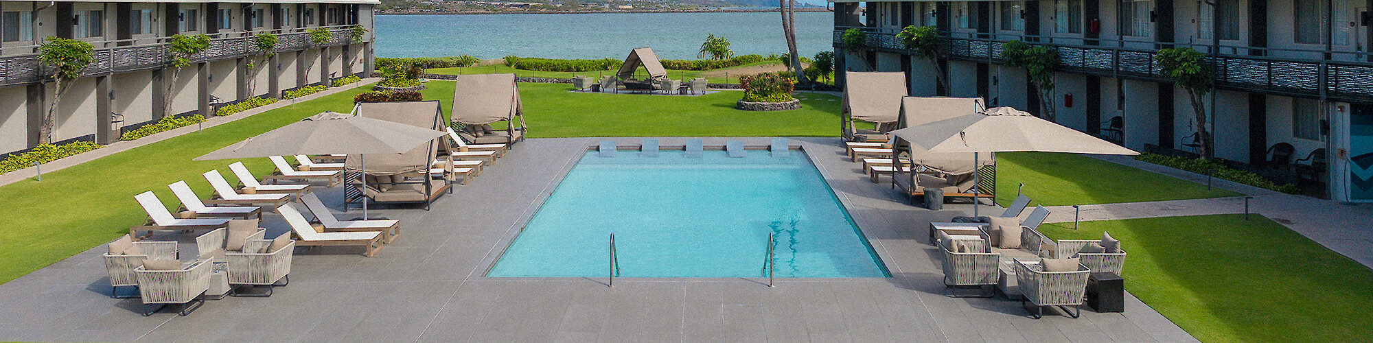 A serene outdoor pool area surrounded by lounge chairs and palm trees, with buildings and mountains in the background under a partly cloudy sky.