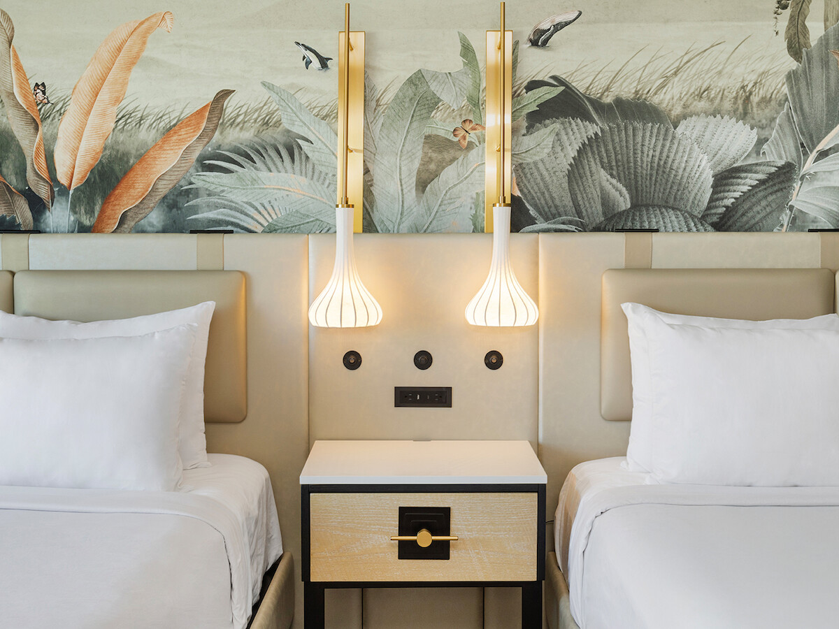 The image shows two beds with white linens, a central nightstand, pendant lights, and a wall with decorative foliage artwork.