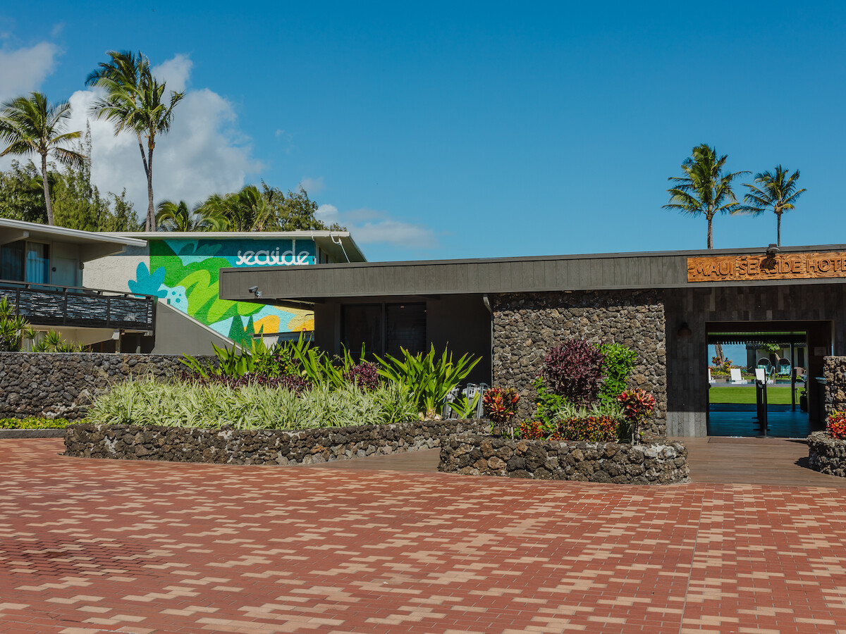 The image shows a modern building with a stone facade, a mural, palm trees, and a large paved area in front under a clear blue sky.