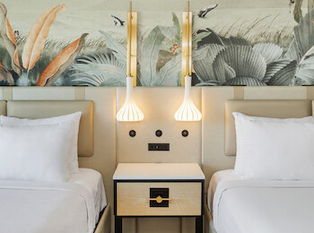 Two neatly made beds with white linens, separated by a nightstand. Above them is a wall mural with botanical patterns and two pendant lights.