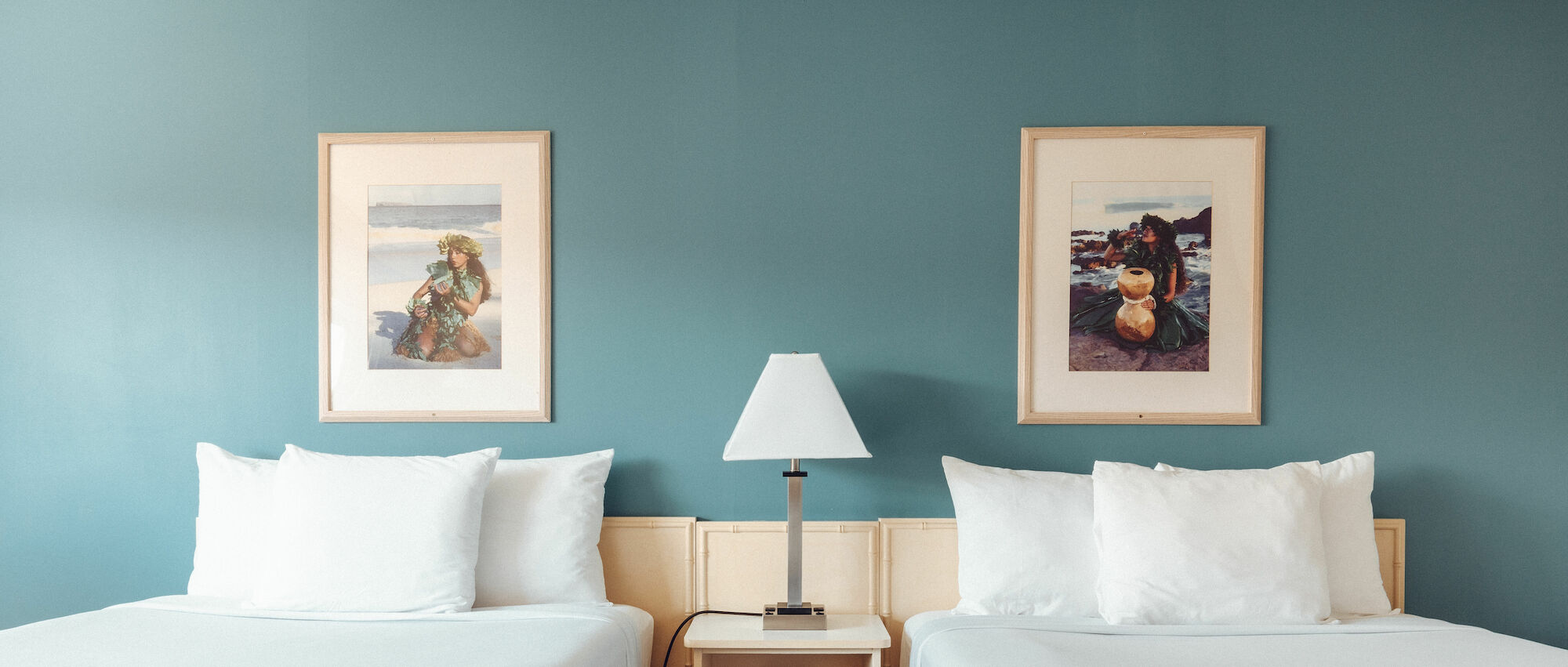 The image shows two twin beds with white bedding, a nightstand with a lamp between them, and two framed pictures above, all set against a teal wall.