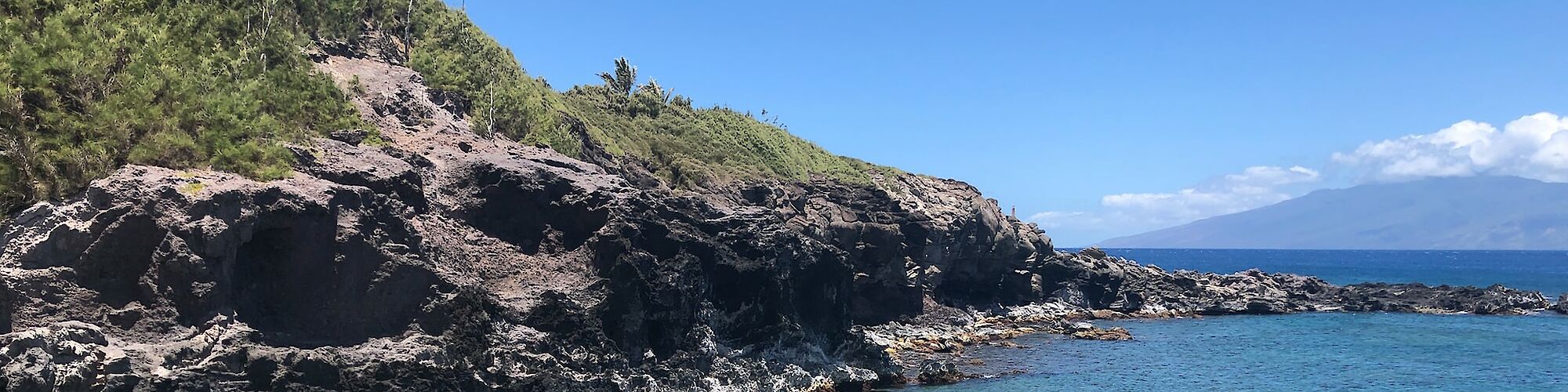 A rocky shoreline with clear blue water, surrounded by rugged cliffs and green vegetation under a bright blue sky with scattered clouds.