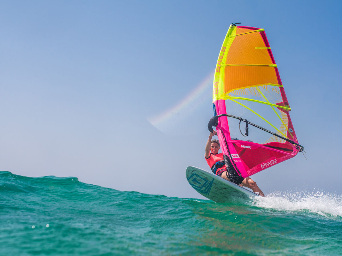 A person is windsurfing on the ocean with colorful sails under a clear blue sky, creating a dynamic and vibrant scene.