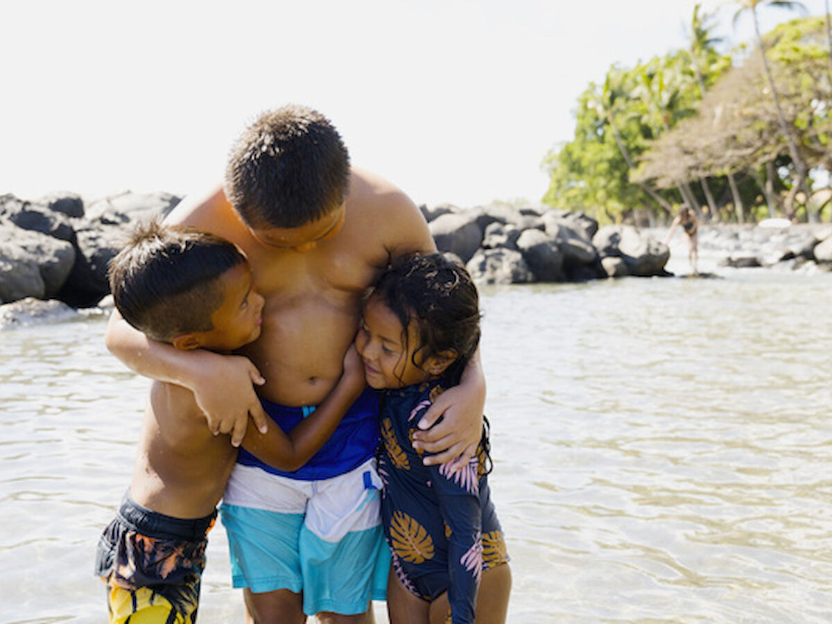 Three children are hugging and smiling while standing in shallow water at a rocky beach with trees in the background.