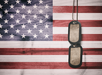 A pair of military dog tags hangs in front of the U.S. flag, suggesting themes of patriotism and military service.