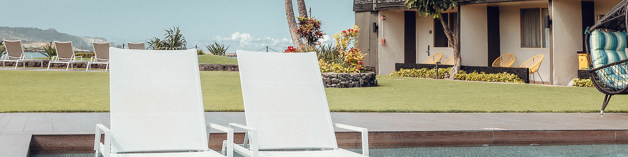 The image shows two white lounge chairs by a pool, with a building and palm trees in the background, under a clear sky.