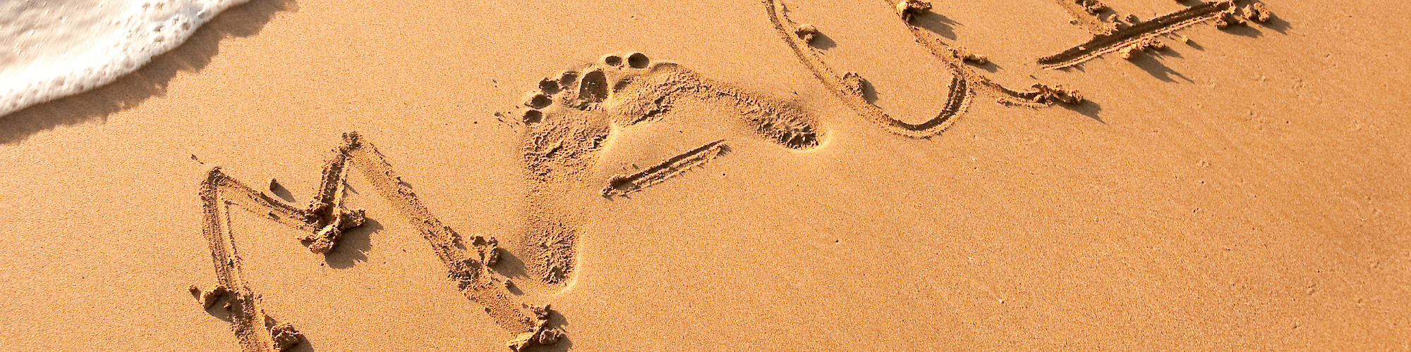 The image shows the word "MAUI" written in the sand on a beach with waves approaching the text.