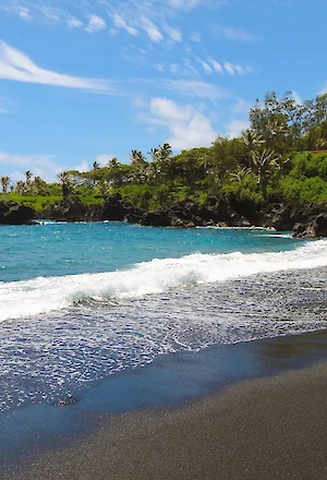 A scenic view of a beach with black sand, turquoise water, and lush green cliffs under a blue sky with white clouds.